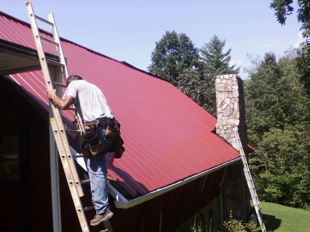 Roofer on a ladder beside a red metal roof and stone chimney — Soffit & Fascia Repair by Roofing Brothers in North Judson
